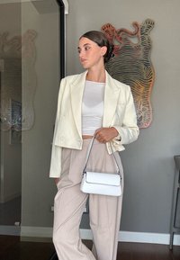 Blazer van ivoorkleurige stof met dubbele knopen, gecombineerd met een aansluitend witte top en lichtbeige wijde broekspijpen, terwijl ze een kleine witte handtas vasthoudt.