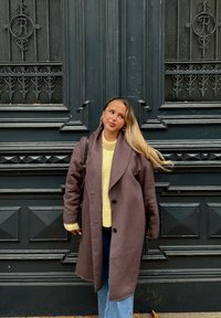 Brun ullkappa med överdimensionerade slag, lagd över en gul stickad tröja. Bärs med blå vida jeans, mot en dekorerad svart vägg.