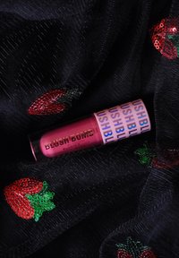 Blush liquide dans un tube rose avec « BLUSH BOMB » en or. Posé sur un tissu noir avec des motifs de fraises en sequins rouges et verts.