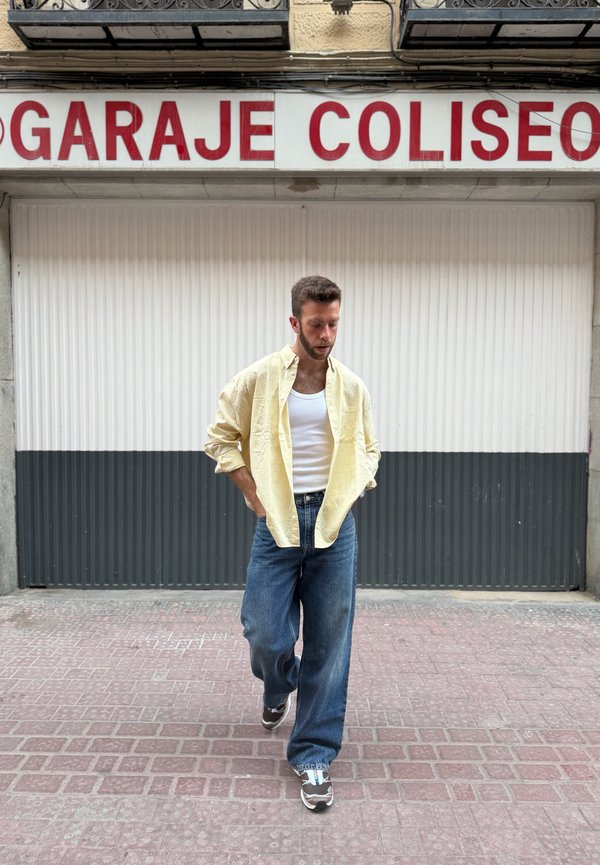 Mann in gelbem Hemd, weißem Tanktop und blauer Jeans, der auf einem gepflasterten Bürgersteig vor einem geschlossenen Garagentor mit dem Schild "GARAJE COLISEO" geht.