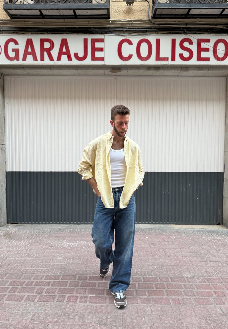 Homme en chemise jaune, débardeur blanc et jean bleu marche sur un trottoir en briques devant une porte de garage fermée avec un panneau "GARAJE COLISEO".