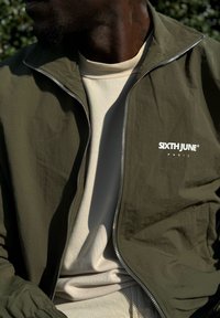 Ελαφριά ελιά πράσινη ζακέτα με φερμουάρ και λευκό λογότυπο στο στήθος, φορεμένη πάνω από ένα κρεμ χρώμα μπλούζα με λαιμόκοψη. Λείος υφασμάτινος τόνος.