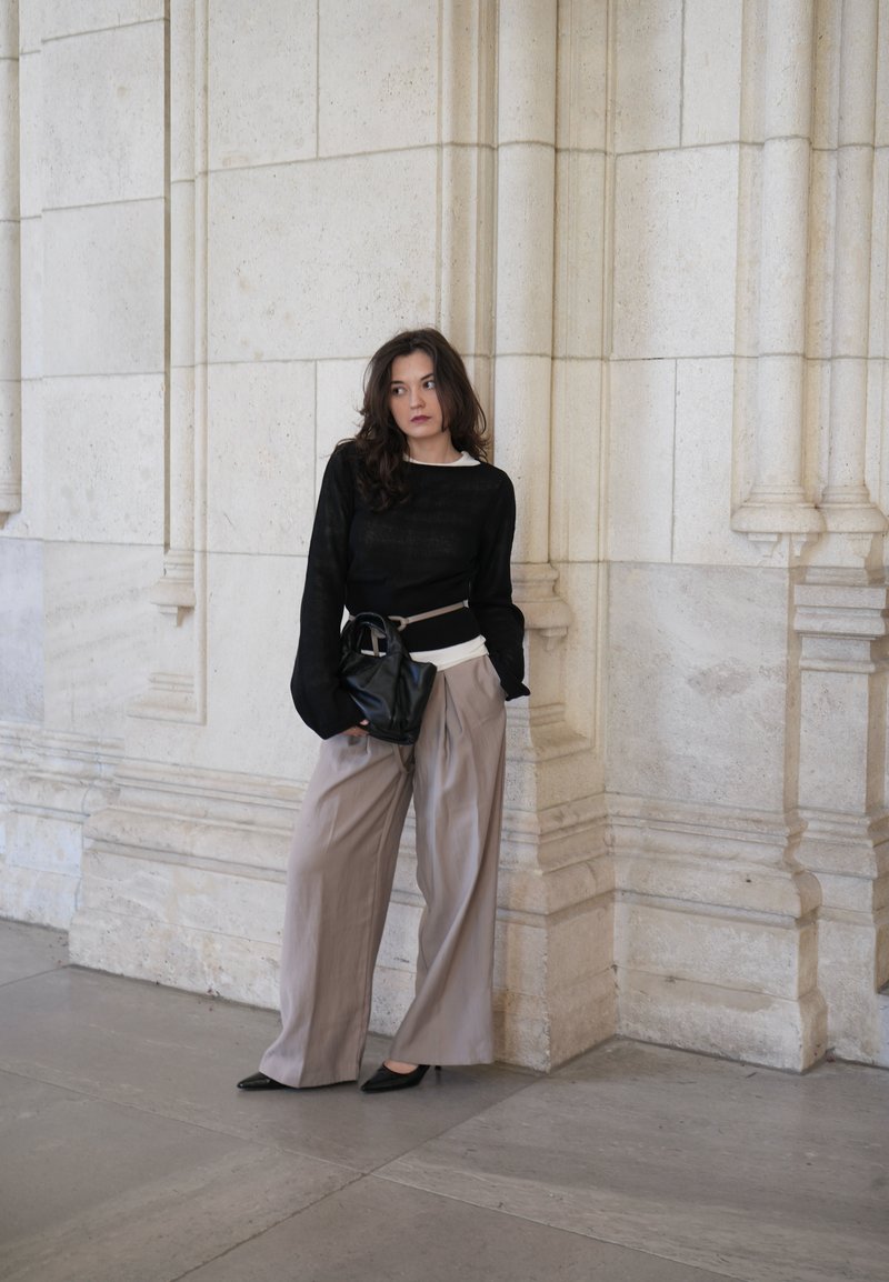 Woman in wide beige trousers, black jumper, and heels standing against a light stone wall with architectural details, holding a black bag at her waist.