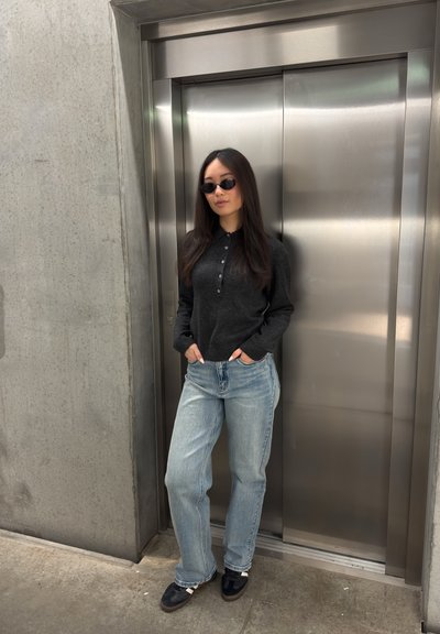 Mujer con gafas de sol, suéter oscuro y vaqueros claros, de pie con las manos en los bolsillos frente a puertas cerradas de ascensor de acero inoxidable.