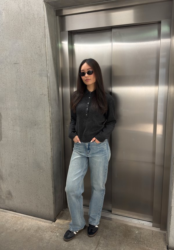 Mujer con gafas de sol, suéter oscuro y vaqueros claros, de pie con las manos en los bolsillos frente a puertas cerradas de ascensor de acero inoxidable.
