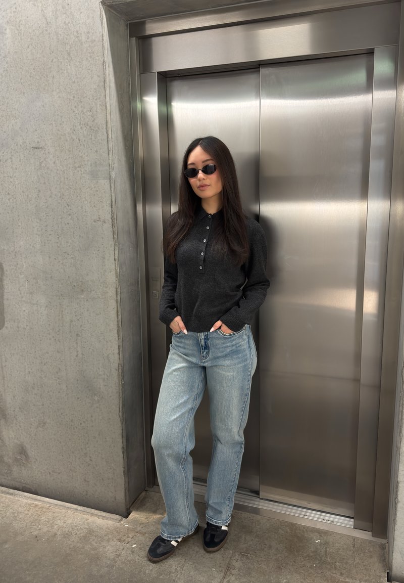 Woman wearing sunglasses, dark sweater, and light jeans stands with hands in pockets in front of closed stainless steel elevator doors.