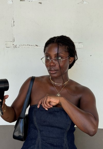 Top negro de mezclilla sin tirantes con un detalle de costura, gafas transparentes, collares de oro en capas, sosteniendo un dispositivo portátil negro, fondo neutro.