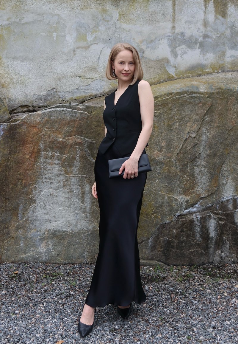 Femme aux cheveux blonds portant une robe noire sans manches et tenant une pochette noire, debout sur du gravier devant un mur en pierre.