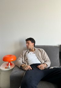 Πορτοκαλί, φωτιστικό σε σχήμα ντόνατ σε μια υφασμένη, ανοιχτόχρωμη πλευρική τραπεζάκι; άτομο με μπεζ κασκόλ κάθεται σε έναν γκρι καναπέ, κοιτώντας στο πλάι.