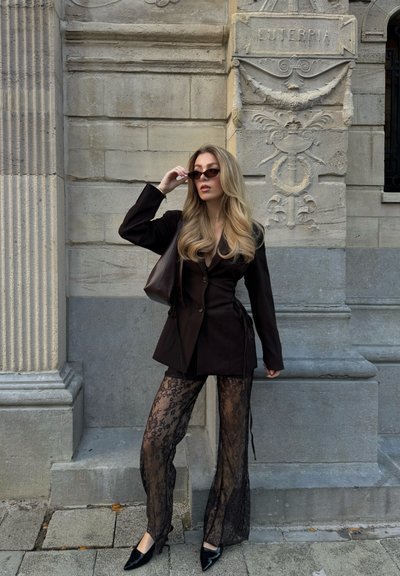 Mujer con blazer negro, pantalones de encaje y zapatos de punta ajustándose las gafas de sol, de pie frente a una fachada de piedra ornamentada.