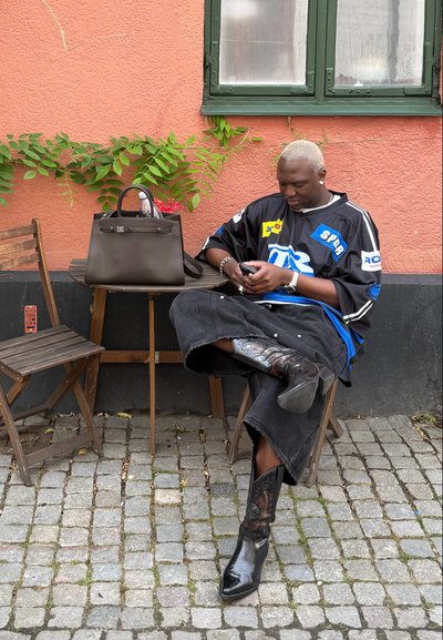 falda de mezclilla negra, camiseta gráfica oversized en negro y azul, botas de vaquero negras y brillantes con diseño intrincado, y un bolso marrón sobre una mesa de madera.