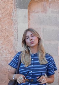 Koszula z krótkim rękawem w niebieskie paski, z guzikami i kołnierzykiem. Osoba trzyma okulary przeciwsłoneczne, ma na sobie pierścionki i długie, falujące blond włosy.