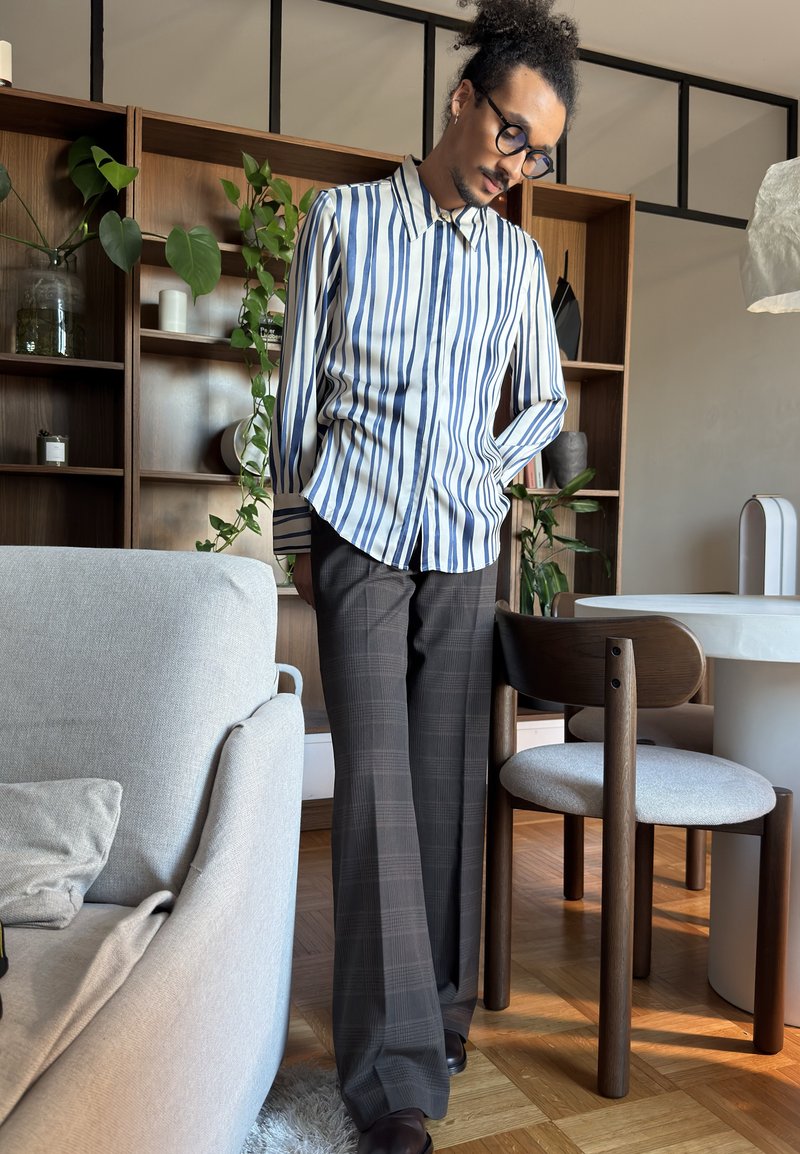 Man som bär glasögon, randig skjorta och rutiga byxor står nära en trästol i ett vardagsrum med växter och hyllor.