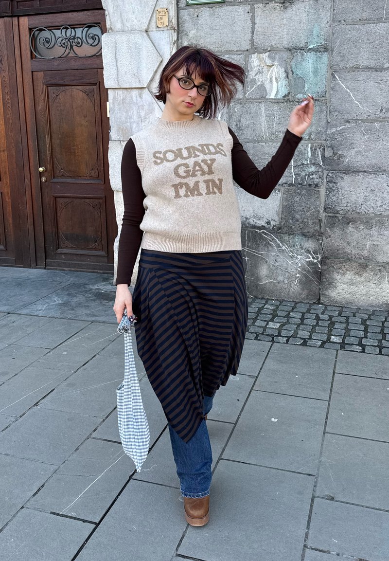 Personne aux cheveux courts et lunettes portant un pull sans manches avec l'inscription « SOUNDS GAY I'M IN », jupe rayée sur un jean, tenant un sac à carreaux.