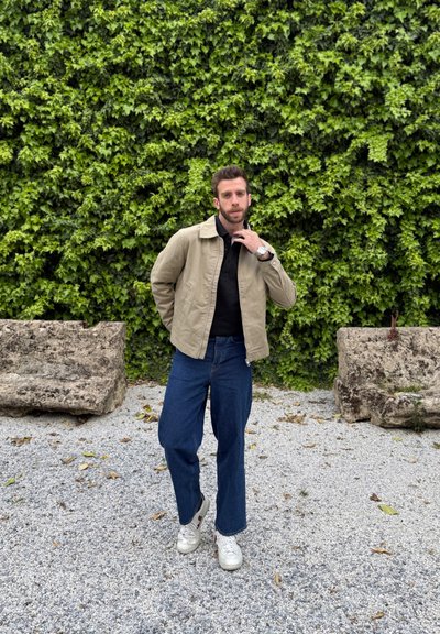 Chaqueta beige clara sobre una camiseta negra, combinada con jeans de pierna ancha azules y zapatillas blancas. De pie frente a una pared de hiedra verde.
