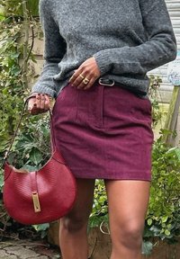 Pullover grigio a maglia, gonna in velluto a coste bordeaux e una borsa rossa in tessuto con una chiusura dorata. Fogliame sullo sfondo.