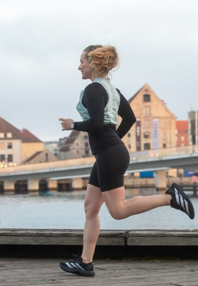Mujer con cabello rojo vestida con ropa deportiva corriendo junto a un río con edificios históricos y un puente al fondo.
