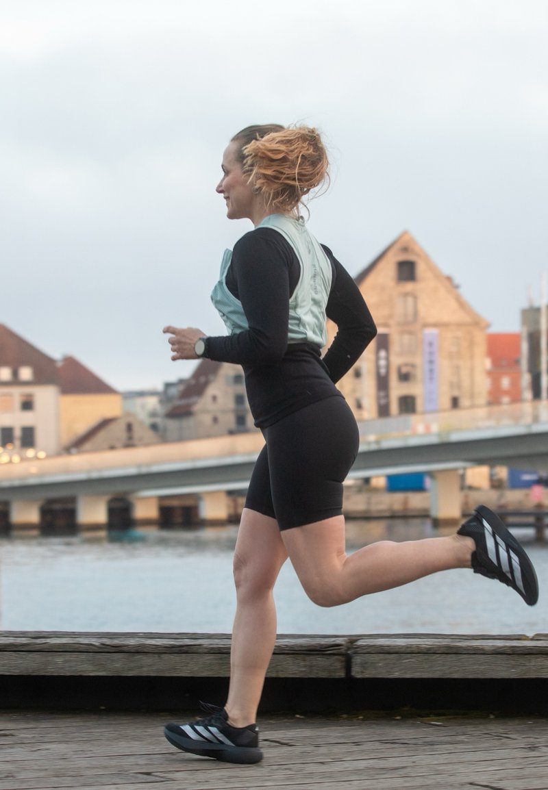 Femme aux cheveux roux en tenue de sport qui fait du jogging le long d'une rivière avec des bâtiments historiques et un pont en arrière-plan.