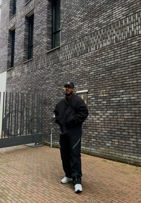Black bomber jacket, black trousers with zippers, white trainers, and a grey cap. Standing against a textured brick wall and metal gate.