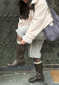 Brown leather knee-high boots featuring buckle accents, a textured surface, and a rubber sole. Styled with grey shorts and a cream coat.