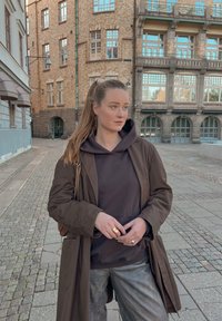 Brun trenchcoat over en mørk hettegenser, kombinert med skinnende grå bukser. Bakgrunnen har teksturerte murbygninger og brosteinsgater.