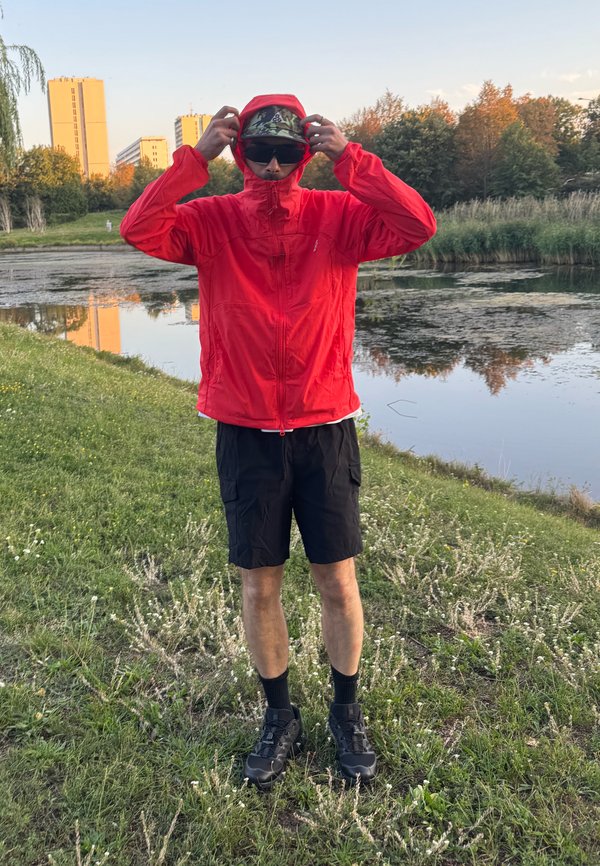 Personne portant une veste rouge, un short noir et des lunettes de soleil, ajustant sa capuche près d'un étang avec des arbres et des bâtiments en arrière-plan.