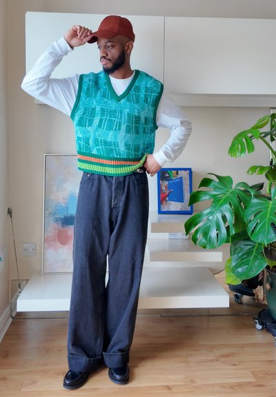 Hombre con gorra roja, chaleco de punto verde con patrón, camisa blanca de manga larga y pantalones anchos negros, de pie en el interior con una planta de interior y estantes flotantes detrás de él.