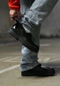 Μαύρα δερμάτινα παπούτσια με χοντρές, υφασμάτινες σόλες, με σχέδιο δέσιμου. Φορεμένα με γαλάζια τζιν που είναι διπλωμένα.