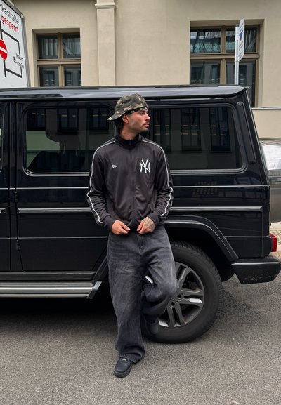 Chaqueta deportiva negra con rayas blancas y logo de NYC, pantalones grises de gran tamaño, gorra tipo bucket de camuflaje; posando junto a un SUV negro sobre el pavimento.