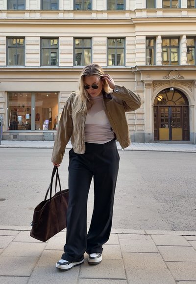 Chaqueta bomber marrón sobre una blusa clara, pantalones amplios negros y zapatillas grises. Lleva un bolso tote marrón oscuro. Entorno urbano.