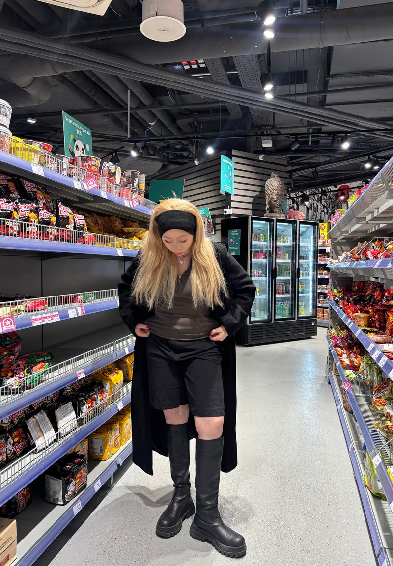 Black knee-high boots, black shorts, and a long coat worn over a sheer top, set in a brightly lit store aisle filled with snack displays.