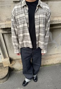 Camisa de flanela aos quadrados em bege e cinza; t-shirt preta por baixo. Jeans escuros com as bainhas dobradas; sapatos pretos completam o conjunto.