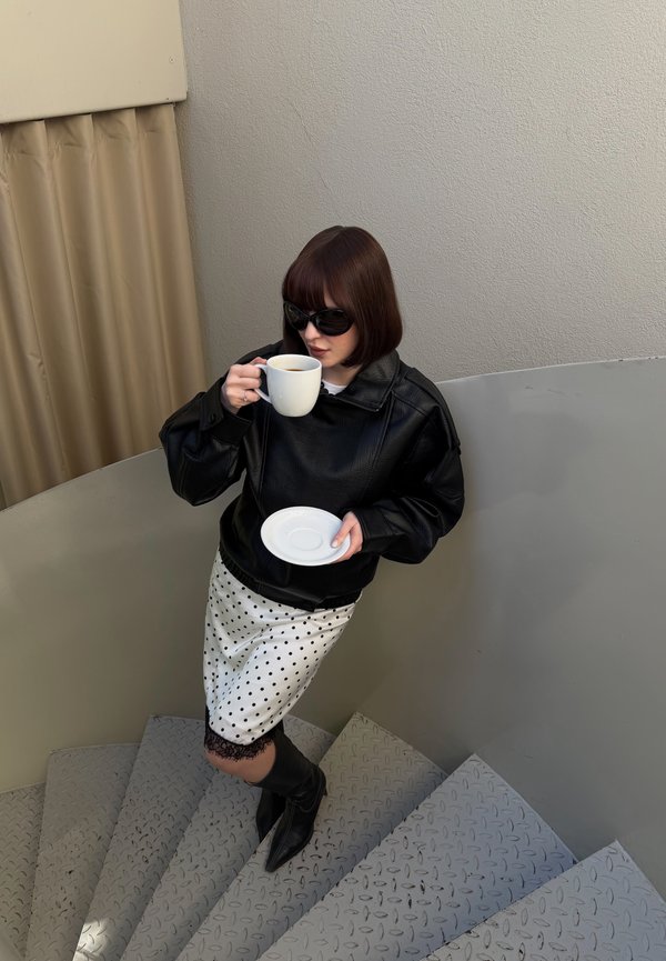 Žena v čiernej koženej bunde, sukni s bodkami a čiernych čižmách pije kávu na kovovom točitom schodisku v interiéri.