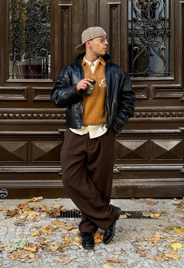 Veste bomber en cuir noir sur un pull beige, chemise à col rayée en dessous, pantalon plissé marron, chaussures noires, tenant une tasse verte.