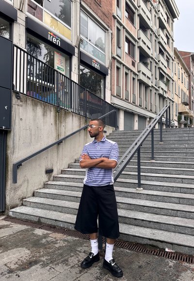 Camisa polo de manga corta a rayas en color púrpura claro y blanco, pantalones cortos negros hasta la rodilla, zapatos negros y calcetines blancos. Escaleras de concreto de fondo.