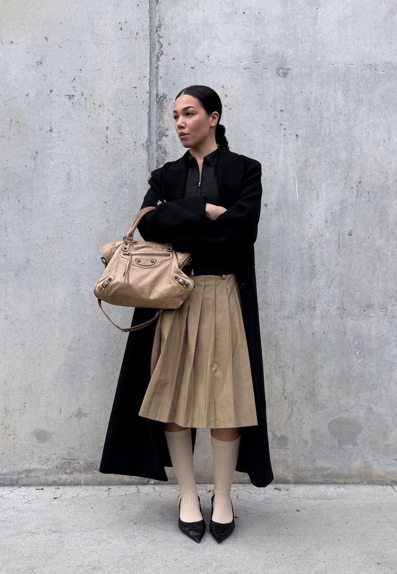 Long black coat, fitted black top, tan pleated skirt, ribbed beige knee-high socks, black pointed-toe heels, and a tan handbag. Concrete background.
