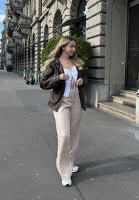 Καφέ δερμάτινο μπουφάν, λευκό cropped top με λεπτομέρεια δέσιμου, μπεζ παντελόνι σε φαρδιά γραμμή και γκρι αθλητικά παπούτσια. Πεζοδρόμιο με πατίνια στο φόντο.