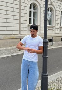 Biała koszulka na krótki rękaw i jasno-niebieskie luźne dżinsy. Stoi obok latarni z słuchawkami w uszach, poprawiając zegarek na nadgarstku.