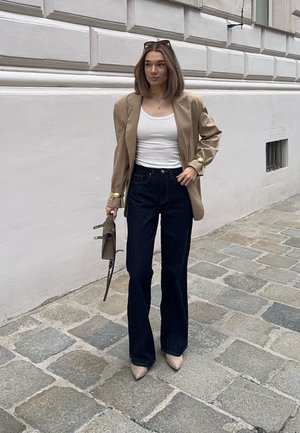 Blazer de satén beige sobre una camiseta blanca, combinado con jeans acampanados de color azul oscuro y tacones de punta nude. Sosteniendo un bolso marrón.