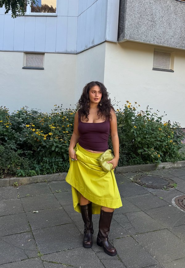 Mujer con cabello largo y rizado que lleva una blusa morada, falda amarilla brillante, botas oscuras hasta la rodilla y sostiene un bolso de mano amarillo a juego al aire libre.
