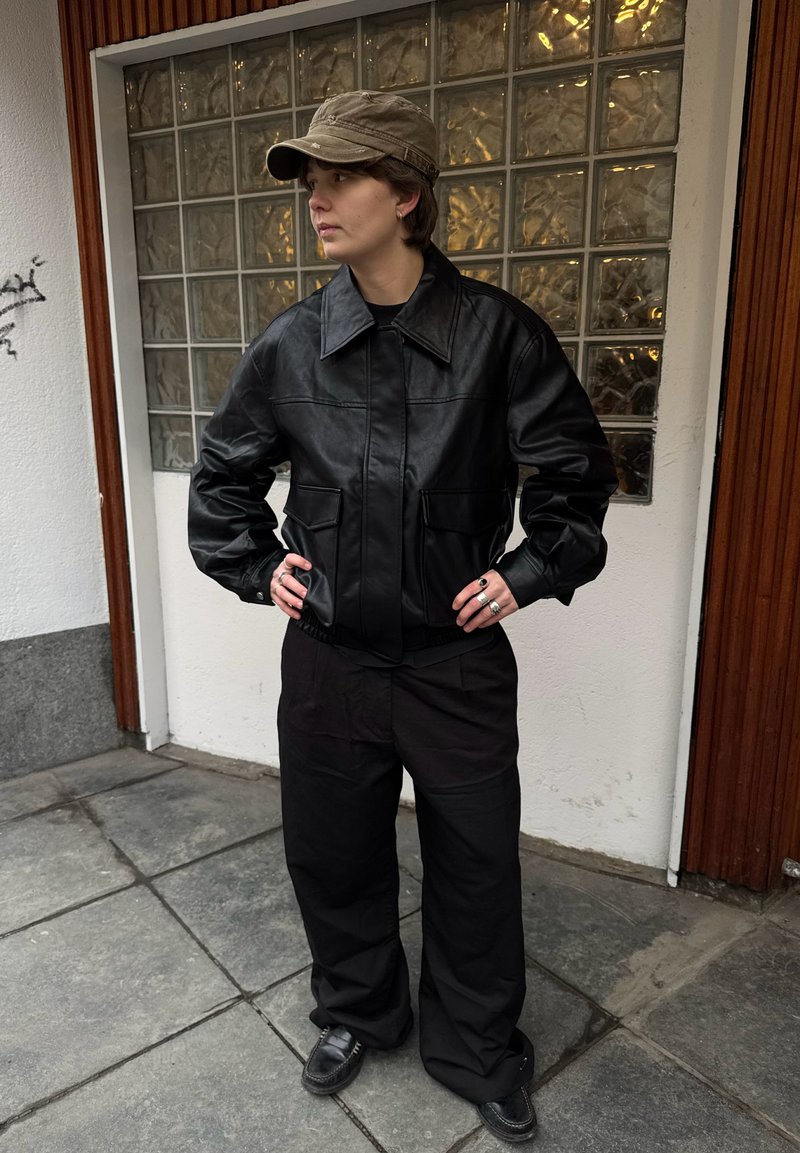 Person in black leather jacket, black wide pants, brown cap, standing with hands on hips against glass block wall and wooden door.
