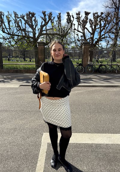 Joven mujer de pie en la calle cerca de árboles sin hojas y bicicletas, lleva una camiseta negra, falda de lunares, medias negras y sostiene un bolso de mano amarillo.