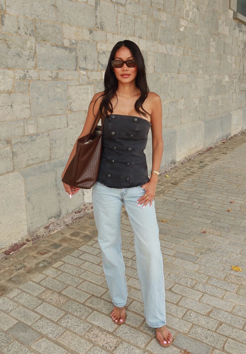Woman with long dark hair wearing a strapless black top, light blue jeans, sunglasses, and carrying a large brown bag standing on a stone pavement.