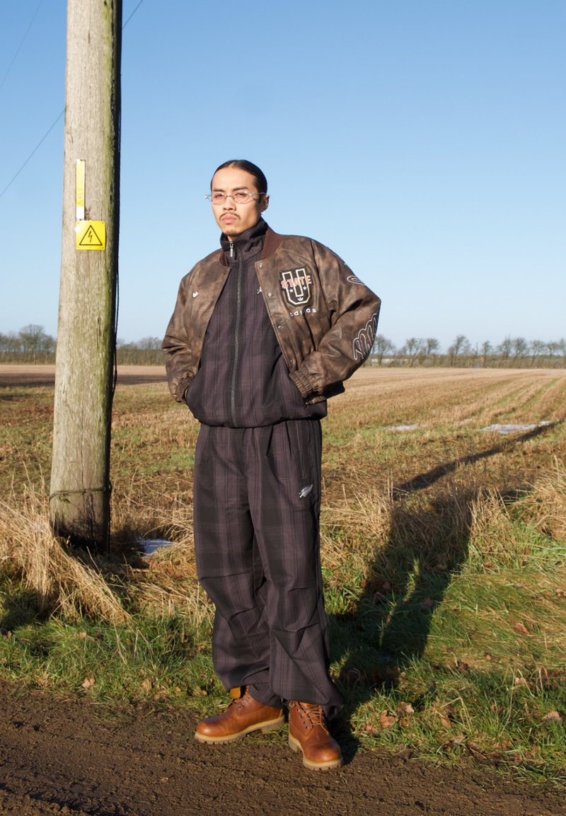 Homme portant une veste en cuir marron, un pantalon à carreaux foncé et des bottes, debout près d'un poteau téléphonique en bois dans un champ ouvert sous un ciel bleu.