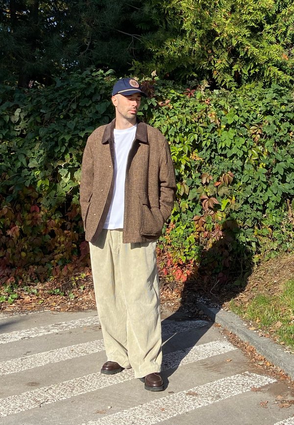Un homme vêtu d’une veste brune, d’une chemise blanche, d’un pantalon beige ample et d’une casquette bleu marine se tient sur un passage pour piétons, à côté de buissons feuillus.