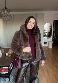 Jeune femme à l'intérieur portant un manteau en fausse fourrure marron, un pull bordeaux, un pantalon en cuir, et tenant un sac à main bordeaux assorti, souriant à la caméra.