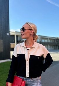 Μαύρο και κρεμ χρώμα-μπλοκαρισμένο κοντό jacket με χρυσά κουμπιά, φορεμένο πάνω από ένα ριγέ μπλουζάκι. Συνδυάζεται με μια κόκκινη τσάντα και γυαλιά ηλίου.