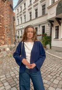 Cardigan bleu marine avec fermeture à boutons et manches larges porté sur un haut blanc. Porté avec un jean ample foncé, sur fond de rue pavée.