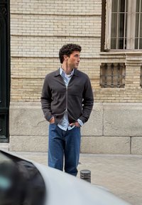 Veste foncée à fermeture éclair avec une poche poitrine et une chemise bleu clair en dessous, associées à un jean bleu ample, sur fond de mur en briques.