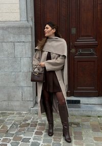 Γυναίκα που στέκεται σε λιθόστρωτο δρόμο, φοράει μπεζ μακρύ παλτό, σκουρόχρωμη πλισέ φούστα, ψηλές καφέ μπότες, κρατώντας σκοτεινό καφέ τσάντα δίπλα σε ξύλινη πόρτα.